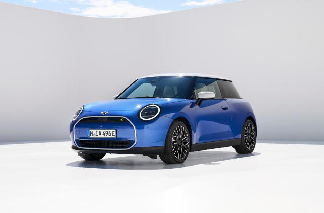 A blue Mini Cooper Electric car is parked in a white space with a light shadow on a white wall behind it.
