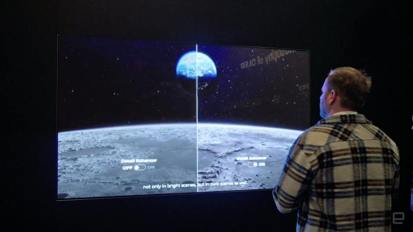 A man stands in front of a large TV display in a dark room. The screen's image is split in half (left and right) to show the differences of picture quality.