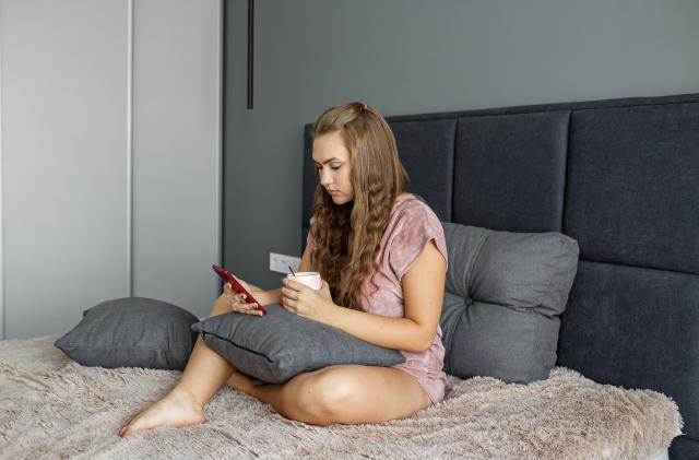 A teenage girl sits in bed and drinks tea. Uses a mobile phone. A woman is texting on social networks using a smartphone.