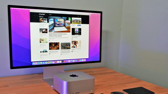 A sparse desktop setup is shown with a Apple Mac Studio and Studio Display sitting on a wooden desktop alongside a keyboard, mouse and touchpage.