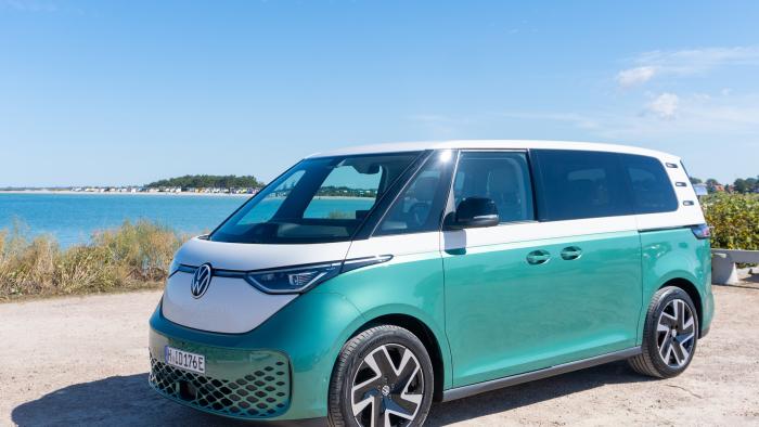 The VW ID.Buzz electric van in teal and white is parked on a dirt or sand area by the water.