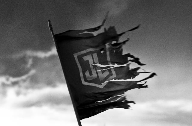 A black and white image showing a torn and tattered Justice League flag flying against a foreboding cloudy sky.