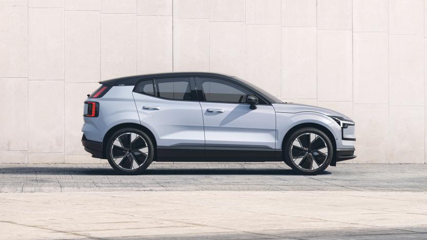 A light grey Volvo EX30 is parked on a grey paved road directly alongside a cream-colored stone wall.