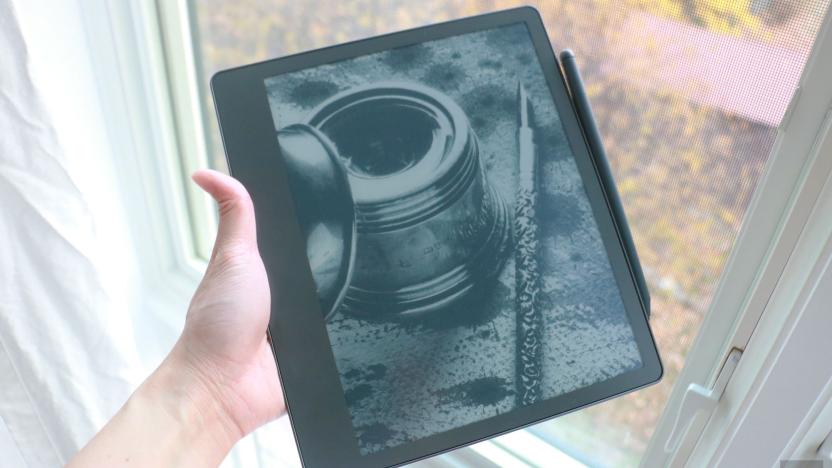 The Amazon Kindle Scribe held in mid-air by a hand gripping its left side, with a pen attached to the right side. In the background is a window with white curtains.