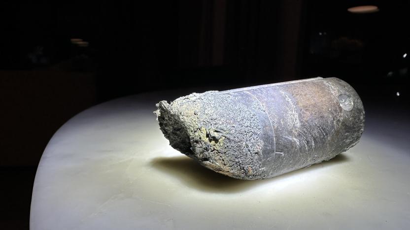 Photo of a stanchion from an ISS space dump. The object, a gray-ish cylinder with crumbled / burned edges, sits on a white table with black background.