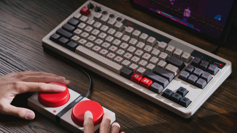 Product lifestyle photo of the 8BitDo Retro Mechanical Keyboard. The keyboard has Nintendo NES-inspired colors with deep (mechanical) keys. A pair of hands mash the two big red Super Buttons wired in front of it.