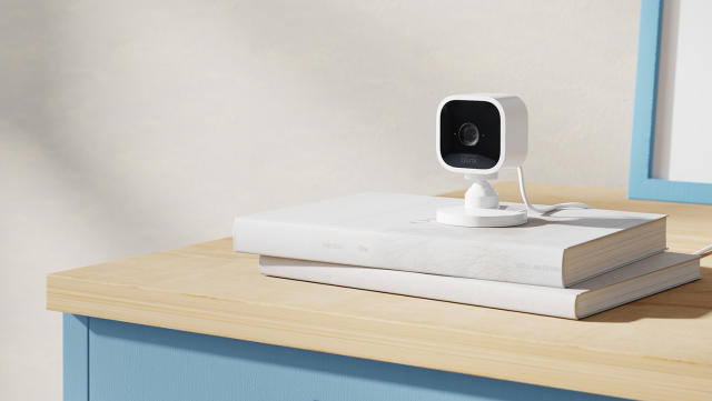 The black and white Blink Mini security camera sits on a pile of books on a wooden cabinet. 