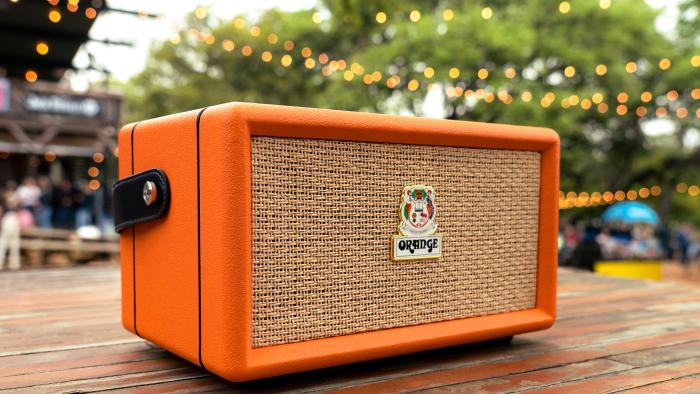 The Orange Box portable Bluetooth speaker sits on a wooden picnic table with string lights overhead, with a crowded outdoor event space and trees blurred in the background.