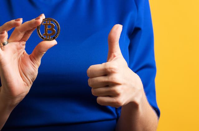 Faceless girl holds golden physical bitcoin and shows Okay sign, thumbs up, ok gesture. Female Hand with Cryptocurrency Coin on Blue Yellow Background. Digital Currencies, Cryptocurrencies, Blockchain