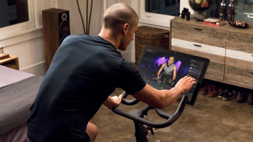 A promotional image of a man in his room riding a Peloton bike with a touchscreen display in front.