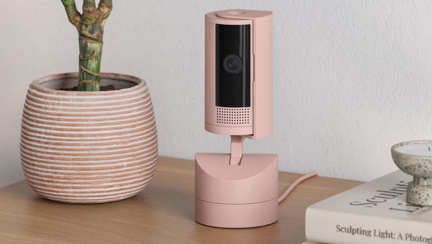 Image of the new Ring indoor pan-tilt camera on a movable base in "blush" which is a fancy word for pink. It is on a table next to a plant pot and some fancy coffee-table books about art that you can't read because there's a dish on top of it.
