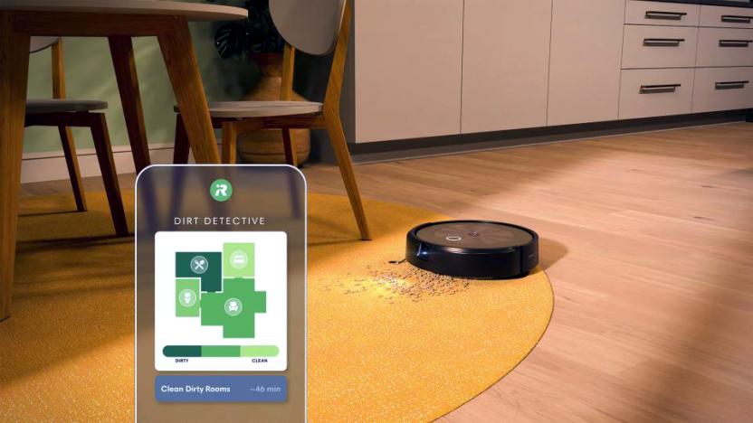 Marketing lifestyle image of the iRobot Roomba Combo J9+. The vacuum moves across a kitchen / dining room floor onto a yellow area rug with dirt and crumbs. A kitchen table and chairs sit next to it with a counter and cabinets behind.