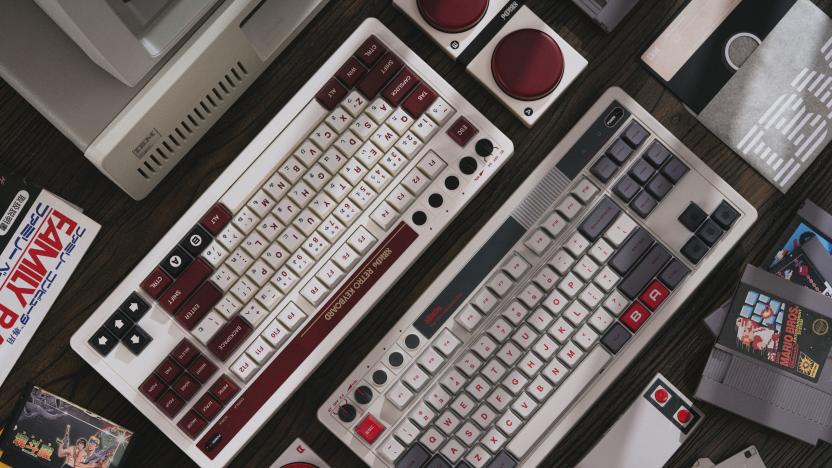 Product photography for the 8BitDo Retro Mechanical Keyboard. Two keyboards (one colored like the NES, the other like Famicom) sit next to each other on a dark desktop. NES cartridges and other 1980s technology nostalgia surround it.