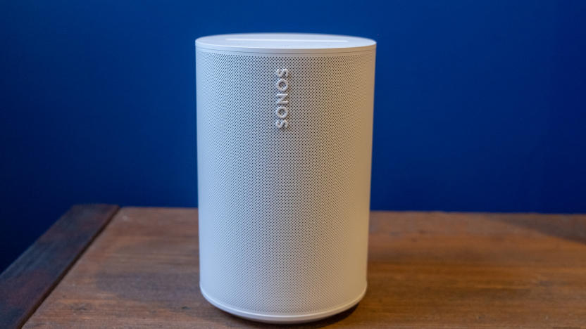 Photo of the Sonos Era 100 smart speaker (white) sitting on a wooden table in front of a purple wall.