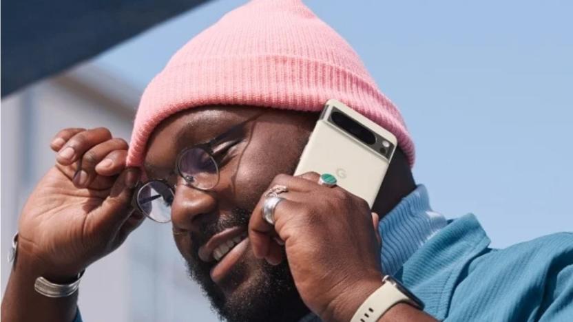 A man wearing a pink beanie and a blue blazer holding a porcelain-colored smartphone against his ear.