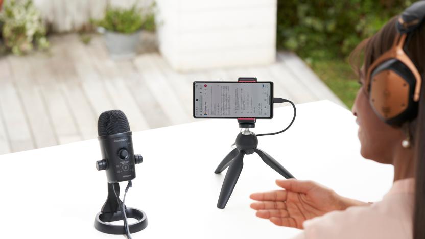 Lifestyle marketing photo of a person (view from behind, only part of them is visible in the right of the frame) talking into a Rode Go:Podcast microphone with a smartphone set up on a mini tripod next to it. White table with a blurred environment behind it.