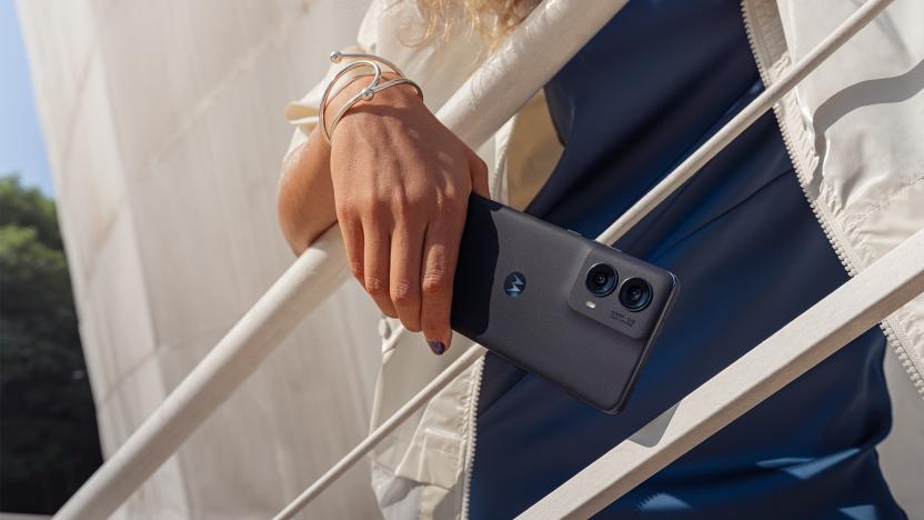 A Person in a white jacket leaning against a white stair railing (outdoors). The 2024 Moto Edge phone is in their hand.