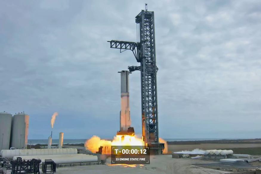 A still image from YouTube of the SpaceX Starship first full static fire test, showing the rocket firing while still attached to its support tower.