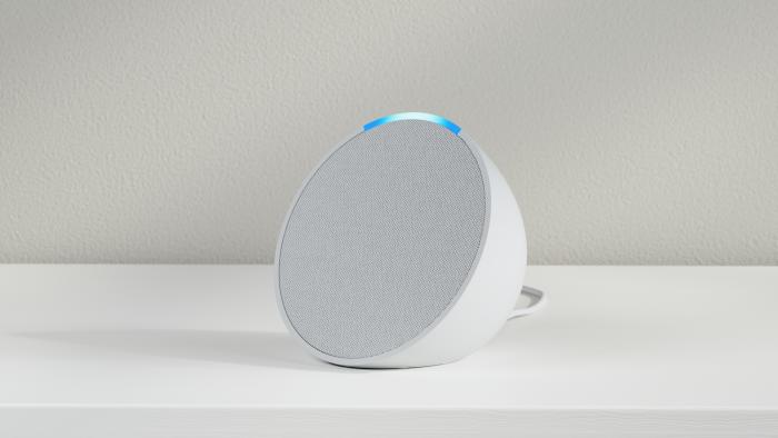 Marketing photo of the semi-spherical Amazon Echo Pop smart speaker. The white speaker sits on a white counter in front of a textured gray wall.