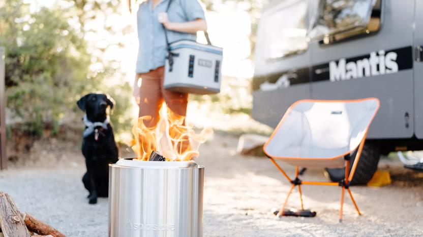 The Solo Stove Ranger fire pit sits on the ground outside with a fire lit inside of it. There is a camper, a camp chair, a black dog and a person carrying a cooler in the background. 