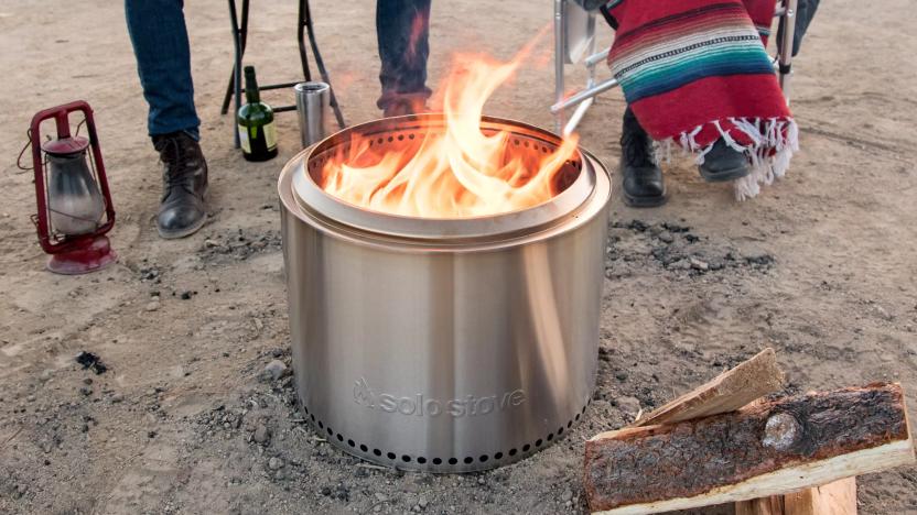 The Solo Stove Bonfire burns wood on a sandy ground. Two people sit in folding chairs in the background. One covers their legs with a colorful blanket. 