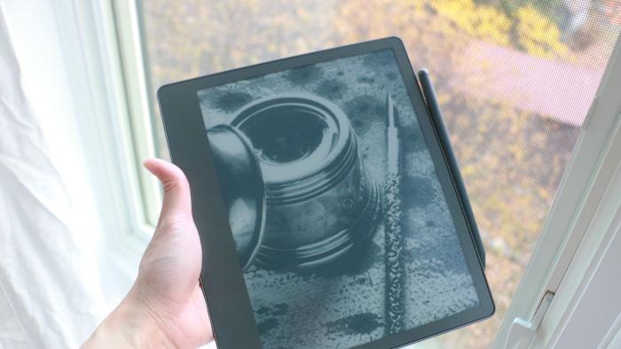 The Amazon Kindle Scribe held in mid-air by a hand gripping its left side, with a pen attached to the right side. In the background is a window with white curtains.