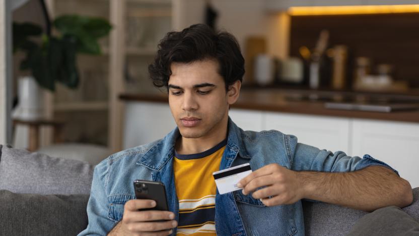 Millennial european man holding plastic credit card in hands, makes purchases on the Internet or uses online banking services sitting on the couch at home