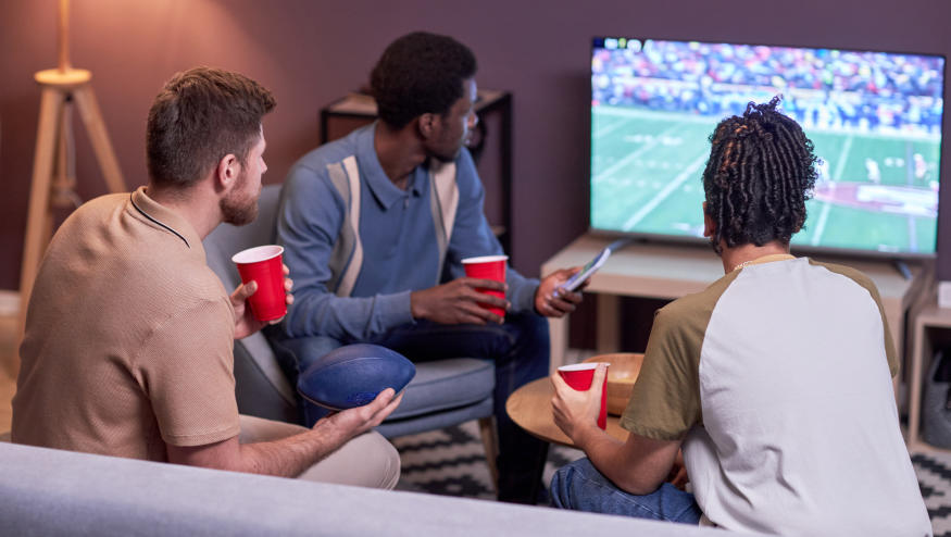 Back view at diverse group of friends watching American football match on TV at home