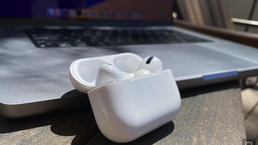 A pair of AirPods in the case sit on a wooden table next to an open MacBook. 