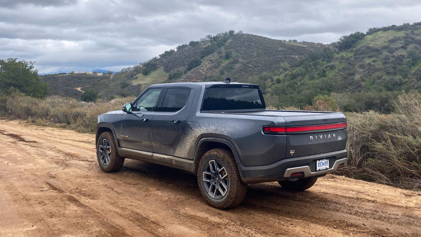 Photo from behind and to the left of the Rivian R1T electric pickup truck. It drives on a dirt road in green hilly terrain.