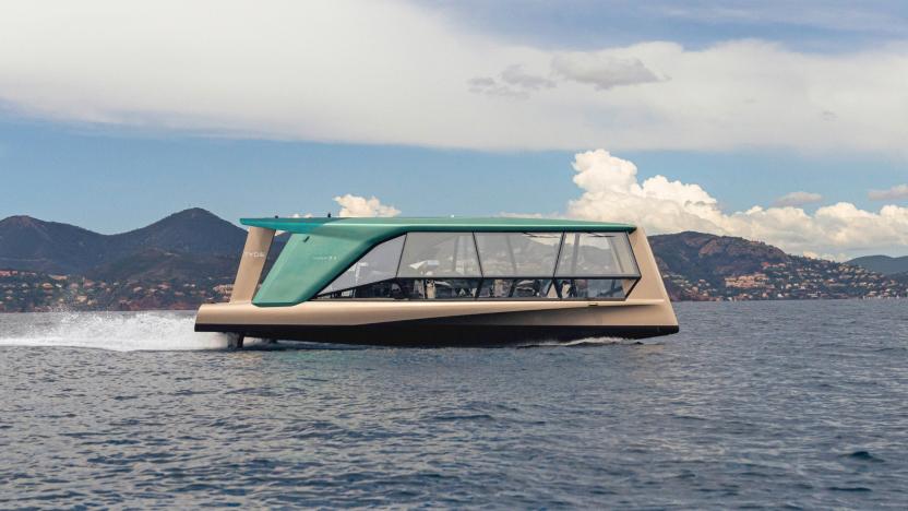 A side view of the glass-walled TYDE/BMW collaborative boat, called the Icon, as it moves across the water with hills and clouds in the distance.