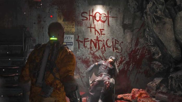 In this still from the video game Callisto Protocol, a character with a screen on his neck is looking at a dead character in the background, with the words "Shoot the tentacles" written in blood on the wall behind him. 