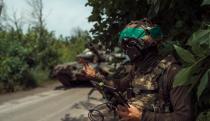 A Ukrainian soldier operating a drone. Forest, next to a street. The solider is holding a tablet with wires as a friendly tank sits behind.