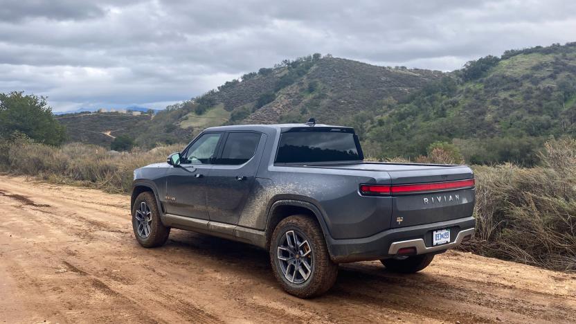 Photo from behind and to the left of the Rivian R1T electric pickup truck. It drives on a dirt road in green hilly terrain.