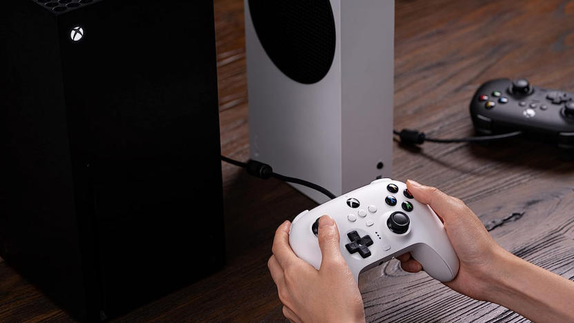 Hands holding a controller with an Xbox on top of a wooden table.