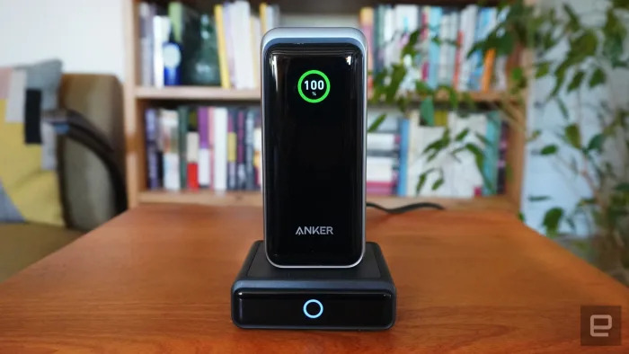 The Anker Prime power bank sits on its charging dock atop a wooden table. There are books and plants in the background. 