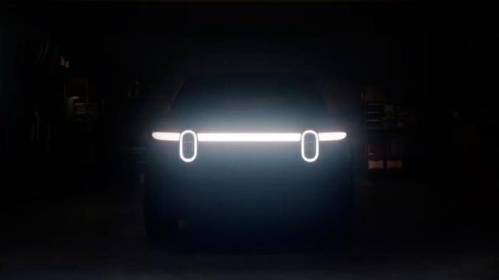 Teaser image for the Rivian R2 EV. Two vertically oval headlights along a sleek horizontal light bar hint at the front of the vehicle. Otherwise nearly black image of a garage.
