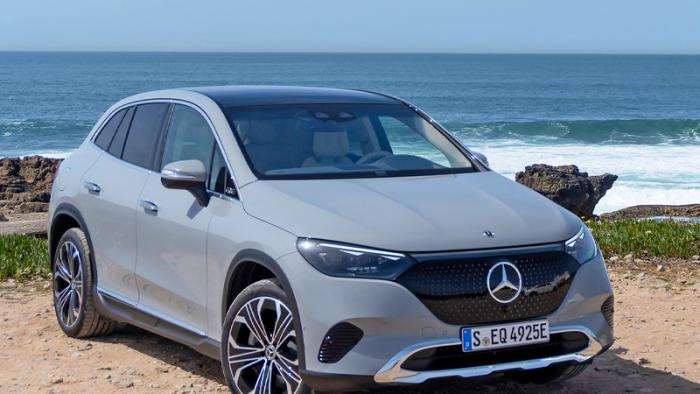 A grey Mercedes EQE SUV is parked on a rocky shore with rock outcroppings and waves close at hand in the background.