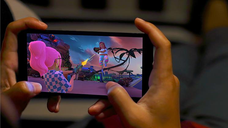 Hands holding a phone with a game on screen. The game shows a pink-haired character shooting at another character in the background.