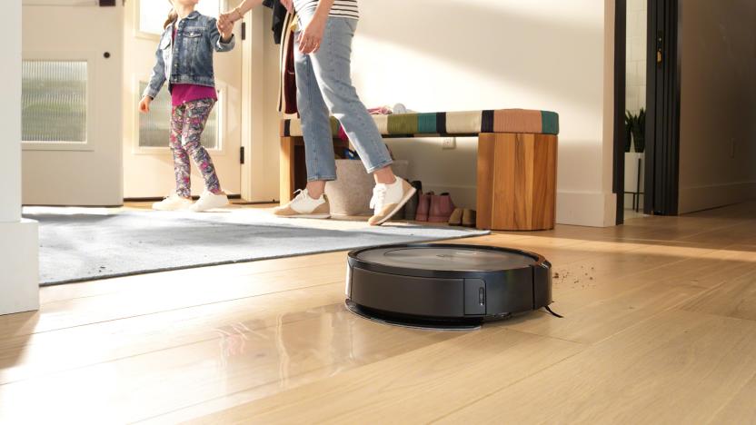 Marketing photo for the iRobot Roomba j5+. The robot vacuum rolls across a family's hardwood floor. In the background, a mother holds a daughter’s hand as they head out the front door.