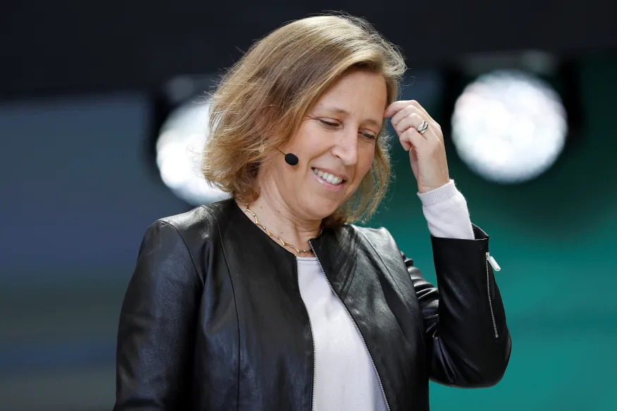 Uma foto de Susan Wojcicki, uma mulher branca, loira e de meia idade, usando um blazer de couro preto, uma camiseta branca e um microfone posicionado na orelha esquerda. O fundo está desfocado e aparenta ter dois holofotes voltados para ela. 