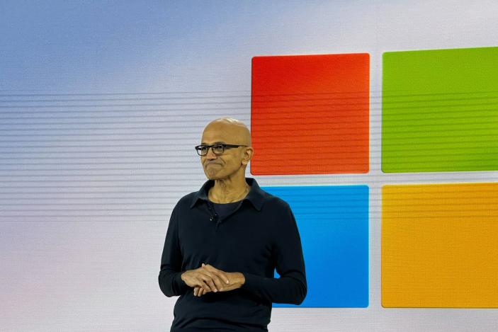 Satya Nadella on stage with the Windows logo on a screen behind him.