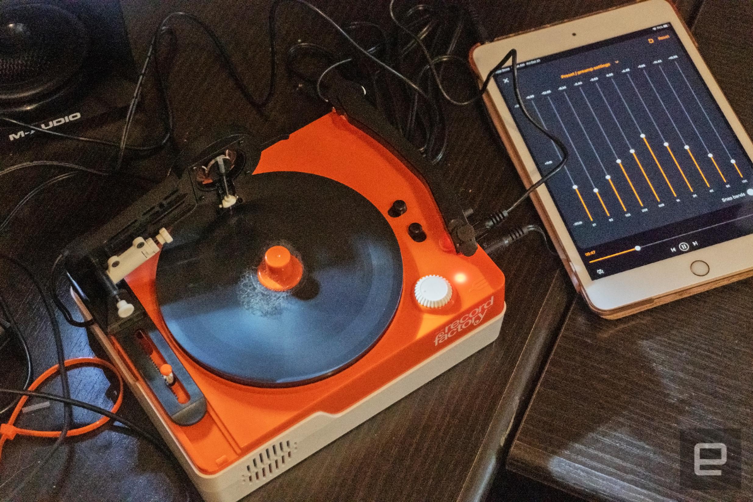 The Teenage Engineering Record Factory vinyl cutter in orange and white seen on a table cluttered with cables, audio gear and speakers.