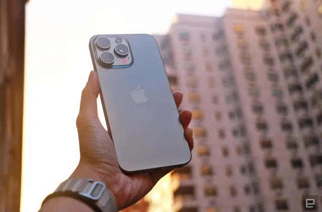 The iPhone 15 Pro Max held up in mid-air with a New York building in the background while the sun sets.   