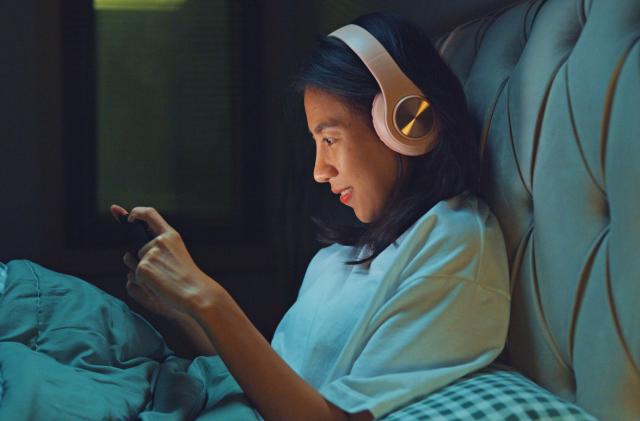 Young Asia girl media addiction on bed wear headphone laugh watch favorite movie in streaming online VOD, video on demand in home at night. Insomnia, Cybersickness, Nomophobia, sleep disorder concept.