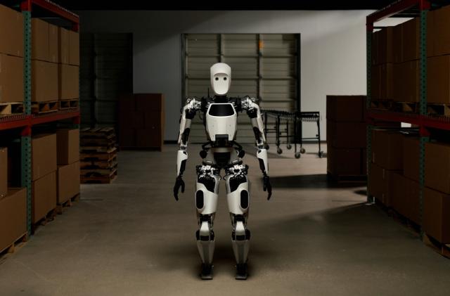 A white and black android stands in a shadowy warehouse corridor, facing the camera.