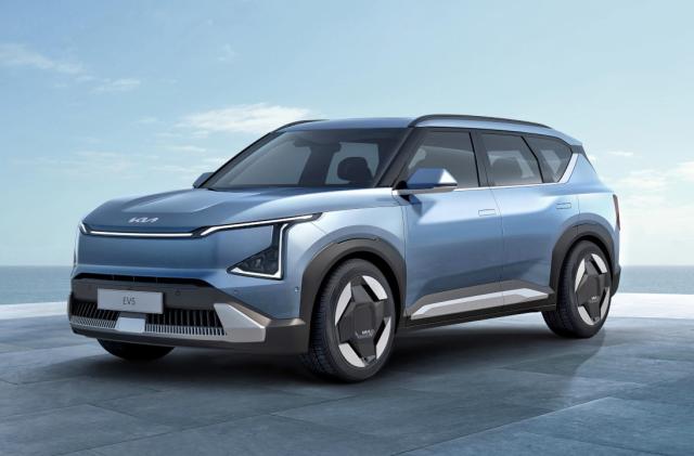 A light blue Kia EV5 SUV is parked on a floor of large tile slabs with the ocean and a blue sky in the background.