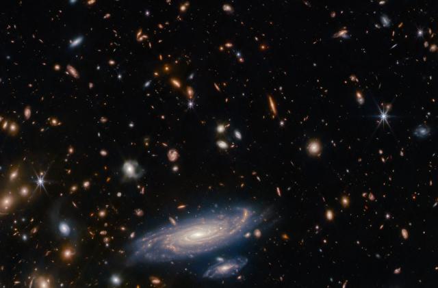 Image that was taken with the James Webb Space Telescope showing a spiral galaxy at the bottom center, surrounded by more distant galaxies (appearing as blobs) and individual stars.