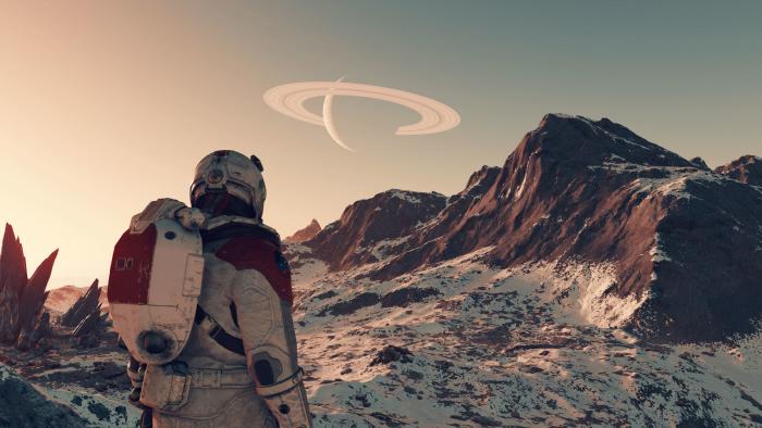 Media image (screenshot) from the game "Starfield." A person in an astronaut suit stands on a planet with reddish-brown mountains. A ringed Saturn-like planet is visible in the daytime sky.