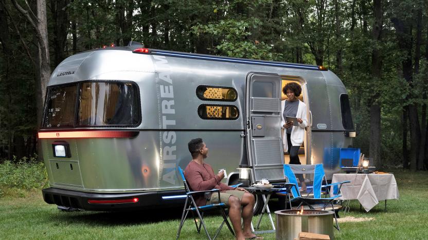 The Airstream eStream electric concept camper parked on the grass by a tree line with a couple sitting outside of it in camping chairs.